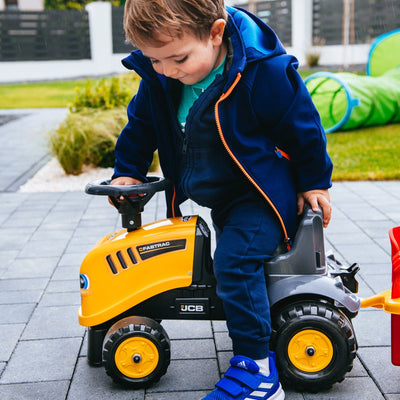 A young child in a blue outfit sits outdoors on a paved surface riding the TP Toys Falk Baby JCB Ride-On Tractor with Trailer, Rake & Shovel.