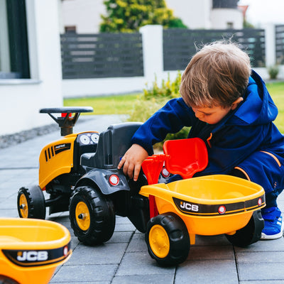 A young child in a blue outfit plays outdoors on a paved surface with the TP Toys Falk Baby JCB Ride-On Tractor with Trailer, Rake & Shovel.