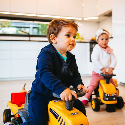 Two children ride TP Toys Falk Baby JCB Ride-On Tractors with trailer, rake & shovel in a modern kitchen. The boy in a blue tracksuit leads, while the girl dressed in pink with a headband follows closely behind on her tractor.