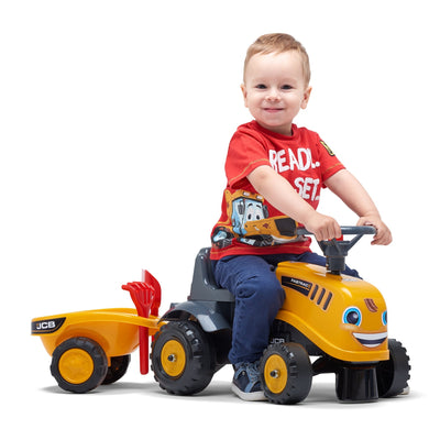 A young child smiles while sitting on the TP Toys Falk Baby JCB Ride-On Tractor with Trailer, featuring a rake and shovel, set against a white background.
