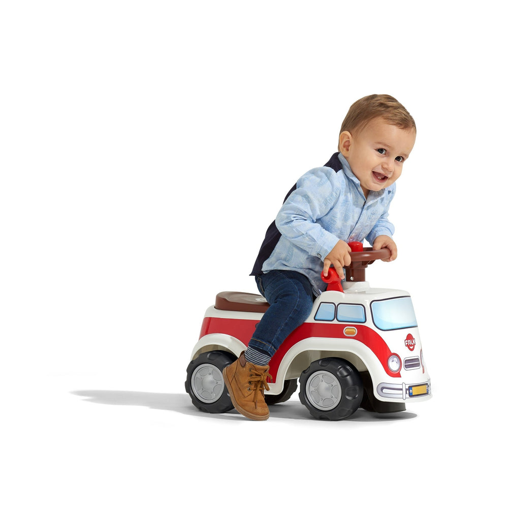 A young child smiles while sitting and riding the TP Toys Falk Retro Combivan Carrier, a red and white toddler ride-on, on a plain white background.