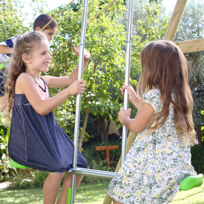 Two young girls enjoy the TP Knightswood Triple Wooden Swing & 8ft Slide Set by TP Toys, sitting and smiling together while a boy stands behind them in a sunny, green garden.