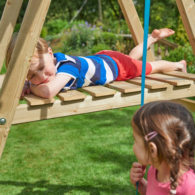 A boy lies on his stomach atop the TP Toys Forest Multiplay Single Wooden Swing & Slide Set while a girl with a hair clip looks up at him from below outdoors.