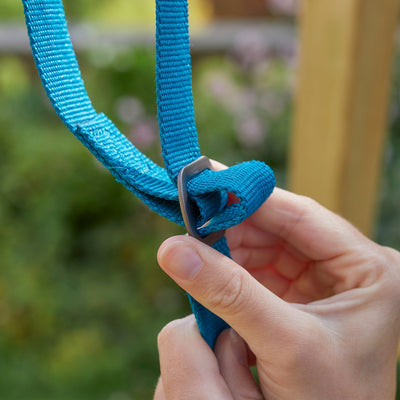 Close-up of hands threading a blue strap through a metal buckle on the TP Toys TP Forest Multiplay Single Wooden Swing & Slide Set - FSC® certified, with a blurred outdoor background.