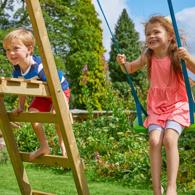 A young boy climbs a TP Toys TP Forest Multiplay Single Wooden Swing & Slide Set while a girl in a pink dress smiles on the swing, enjoying this FSC® certified play frame in a sunny garden.