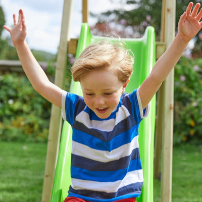 A young boy with blond hair and a striped shirt slides down the wavy slide of the TP Toys Forest Multiplay Single Wooden Swing Set with Roped Rapide Seat, arms raised and smiling.