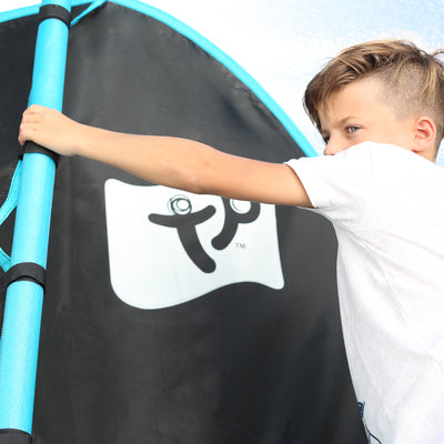 A young boy in a white shirt holds onto a blue padded pole beside a black and blue TP Toys TP Genius® 14ft Round Trampoline featuring a white logo.