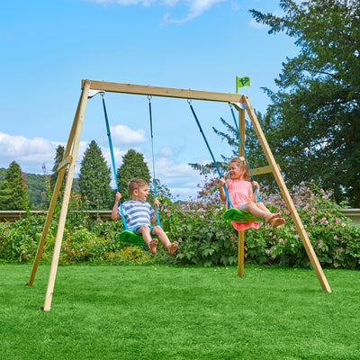 Two young children smile at each other on the TP Toys TP Forest Wooden Double Swing—FSC® certified, surrounded by lush greenery and blooming flowers in the garden.