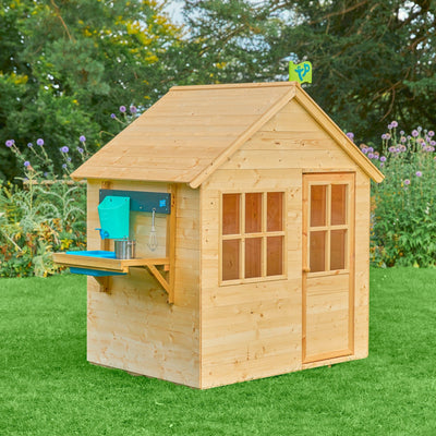 The TP Toys TP Hideaway Wooden Playhouse with Mud Kitchen – FSC® certified – features a pitched roof, two windows, a door, and an attached outdoor kitchen, standing on grass with plants in the background.