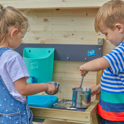 Two young children have fun outdoors with the TP Toys TP Hideaway Wooden Playhouse with Mud Kitchen - FSC® certified, playing with metal pots and utensils at their rustic mud kitchen beside the playhouse.