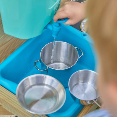 A child pours water from a blue plastic tap into a metal pot at the TP Hideaway Wooden Playhouse with Mud Kitchen by TP Toys, with two more metal dishes in a blue basin nearby.