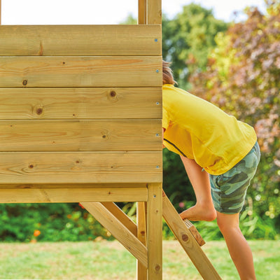 A young child climbs the TP Toys TP Treetops Wooden Tower Playhouse with Toy Box – FSC® certified, surrounded by trees and grass.