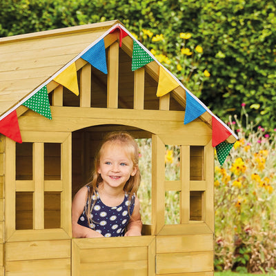 A young girl smiles, looking out the window of the TP Toys Dandelion Cottage Playhouse with Mud Kitchen, Table & Bench and Shutters (FSC® certified), decorated with colorful bunting in a garden.