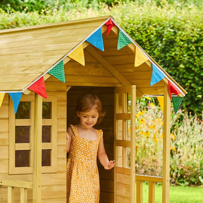 A young child in a yellow dress stands at the doorway of the TP Toys Lavender Cottage Playhouse with Deluxe Mud Kitchen Accessory, FSC® certified. Colorful flags decorate the wooden playhouse, with greenery and flowers in the background.