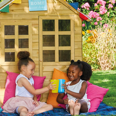 Two children sit on a blanket in front of the TP Toys Foxglove Cottage Playhouse with Nought & Crosses and Basketball Hoop—an FSC® certified playhouse—clinking colorful cups together for imaginative garden fun.