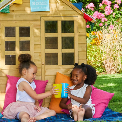 Two young girls smile and clink colorful cups on a blanket before the TP Toys Foxglove Cottage Playhouse with Early Fun Mud Kitchen Accessory and Shutters, made from FSC® certified wood, surrounded by bright pillows and a lush garden.
