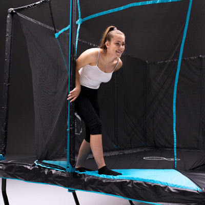 A young woman in athletic wear smiles as she steps onto the TP Toys Infinity 10ft x 14ft Rectangular Trampoline, holding the zip-free entry flap of its safety net.