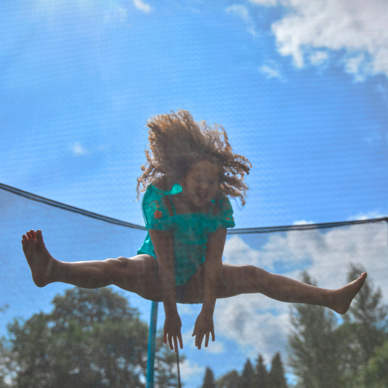 A child with curly hair jumps midair on the TP Toys TP Challenger® 8 ft Trampoline outdoors, legs spread and arms reaching down, while the patented SurroundSafe enclosure keeps them safe under a blue sky with clouds.