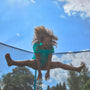 A child with curly hair jumps midair on the TP Toys TP Challenger® 8 ft Trampoline outdoors, legs spread and arms reaching down, while the patented SurroundSafe enclosure keeps them safe under a blue sky with clouds.