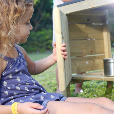 A young child in a blue dress sits outside with the TP Toys TP Junior Chef Wooden Mud Kitchen - FSC® certified. A metal cup on the ledge adds charm to this cozy children’s play kitchen scene.