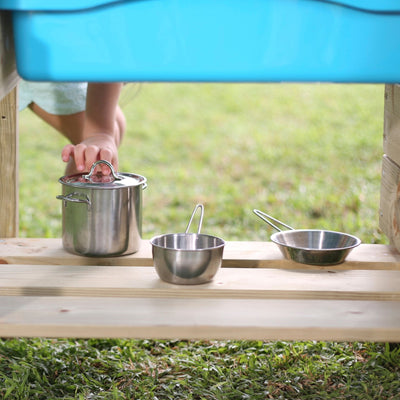 A child reaches for a small metal pot on a wooden shelf in the TP Junior Chef Wooden Mud Kitchen by TP Toys, with two metal measuring cups nearby and grass visible in the background.