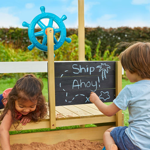 Two young children play on the Ahoy Wooden Playboat by TP Toys, styled as a pirate ship with a blue steering wheel and a chalkboard sign reading "Ship Ahoy!" complete with palm tree and treasure map drawings. FSC® certified wood.