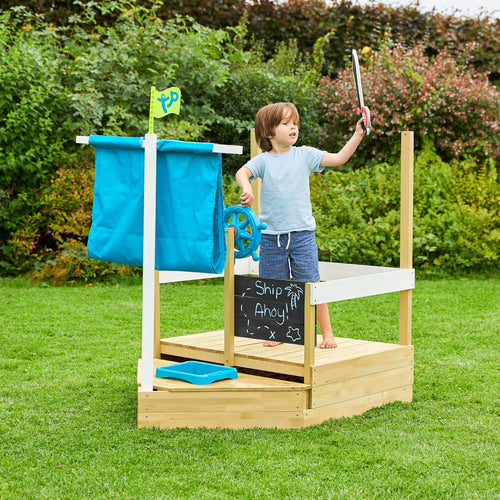A young child stands on the TP Toys Ahoy Wooden Playboat - FSC® certified, featuring a blue sail, steering wheel, and "Ship Ahoy!" sign—perfect for sand and water play in a grassy garden.