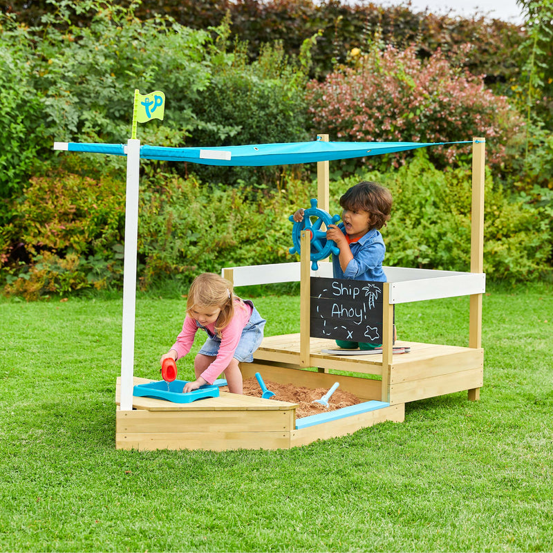 Two young children enjoy imaginative play in the TP Toys TP Ahoy Wooden Playboat with Dig & Explorer Accessory Kit—featuring a steering wheel, chalkboard, and blue canopy—for endless sand and water adventures. FSC® certified.