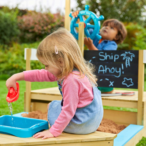 Two young children play on the TP Toys Ahoy Wooden Playboat—one pours water as the other steers the ship’s wheel, imagining a pirate adventure. “Ship Ahoy!” is written on the chalkboard behind their FSC® certified playset.