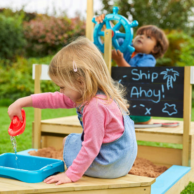 Two children embark on adventures with the TP Toys TP Ahoy Wooden Playboat with Dig & Explorer Accessory Kit—one pours water, the other steers by the “Ship Ahoy!” sign—transforming sand and water play into imaginative fun.