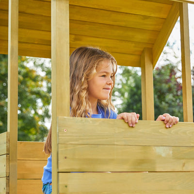 A young girl with long hair stands behind the wooden railing of the TP Toys TP Treehouse Wooden Play Tower with Wavy Slide (FSC® certified), looking to the side. Trees and greenery fill the background.