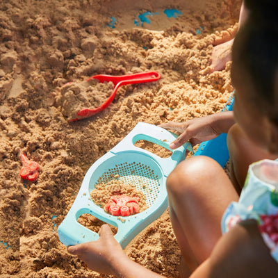 A child enjoys imaginative play on a sunny day with the TP Toys TP Ahoy Wooden Playboat with Dig & Explorer Accessory Kit—featuring a blue sifter, red sand toys, and a red shovel—all FSC® certified.