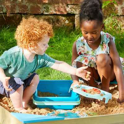 Two young children embark on a wooden play boat adventure outdoors with the TP Ahoy Wooden Playboat and Dig & Explorer Accessory Kit by TP Toys, using the included sand toys to scoop and shape in their sandbox.