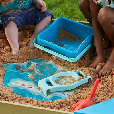 Two children enjoy imaginative play in the TP Toys Ahoy Wooden Playboat with Dig & Explorer Accessory Kit, sifting sand with colorful tools, while grass provides a lush backdrop. The playboat is FSC® certified for sustainability.