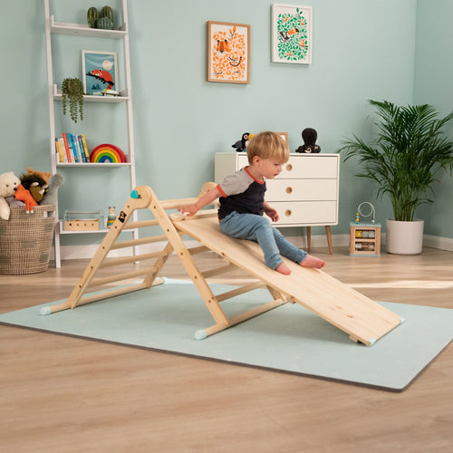A young child sits on the TP Active-Tots Pikler Style Wooden Climbing Bridge and Slide by TP Toys—an FSC® certified Montessori toy—enjoying safe indoor play in a playroom filled with toys, plants, and framed pictures.