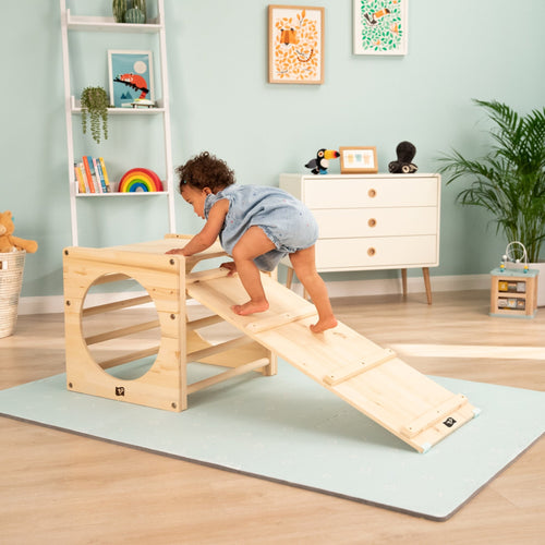A toddler climbs the TP Active-Tots Pikler Style Wooden Climbing Bridge and Slide by TP Toys in a bright, toy-filled room—ideal for indoor play. This FSC® certified product adds fun and style to any space.