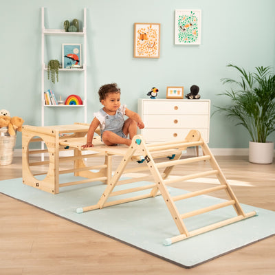 A toddler sits on the TP Toys Active-Tots Pikler Style Wooden Climbing Bridge and Slide (FSC® certified) in a playroom filled with toys, books, plants, and framed art against pastel walls.