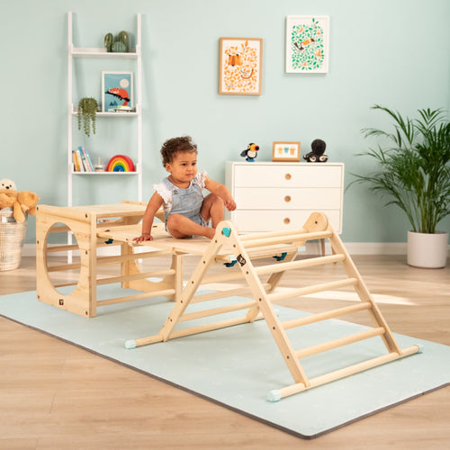A toddler sits on the TP Toys Active-Tots Pikler Style Wooden Climbing Bridge and Slide (FSC® certified) in a playroom filled with toys, books, plants, and framed art against pastel walls.