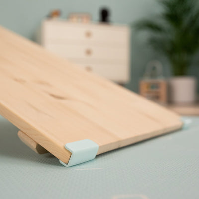 The TP Toys TP Active-Tots Pikler Style Wooden Climbing Bridge and Slide – FSC® certified, features light blue corner brackets and is set up at an angle indoors for active play. A dresser and a plant can be seen blurred in the background.