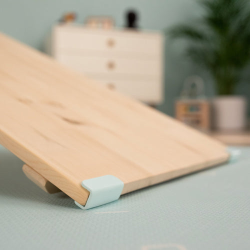 The TP Toys TP Active-Tots Pikler Style Wooden Climbing Bridge and Slide – FSC® certified, features light blue corner brackets and is set up at an angle indoors for active play. A dresser and a plant can be seen blurred in the background.