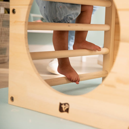 A child climbs barefoot on the TP Toys Active-Tots Pikler Style Two Climbing Cubes & Bridge, carefully stepping on each FSC® certified wooden rung of this Montessori-inspired indoor set.