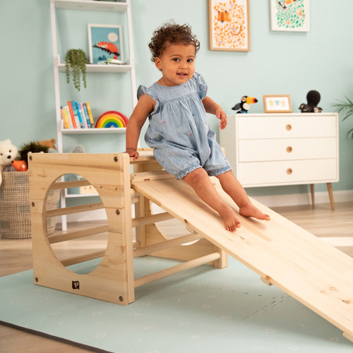 A toddler in a blue romper slides down the TP Toys Active-Tots Pikler Style Two Climbing Cubes & Bridge (FSC® certified) in a playroom with pastel walls, books, toys, and framed art in the background.
