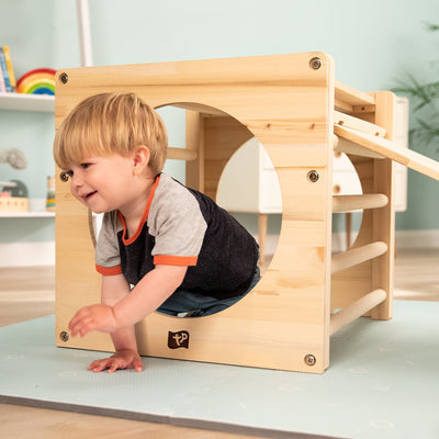 A young child crawls through the TP Active-Tots Pikler Style Wooden Climbing Cube by TP Toys, FSC® certified, creating an engaging indoor play environment surrounded by toys and shelves.