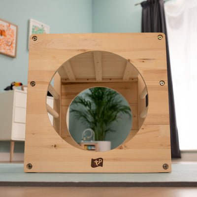 The TP Toys TP Active-Tots Pikler Style Wooden Climbing Cube—FSC® certified—features a large circular opening and sits indoors, with a potted plant and other furniture in the background, creating an inviting play space.