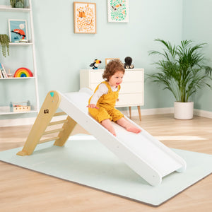 A young child in yellow overalls slides down the TP Toys TP Active-Tots Pikler Style Folding Wooden Slide—an FSC® certified indoor play piece—in a brightly lit, neatly decorated playroom.