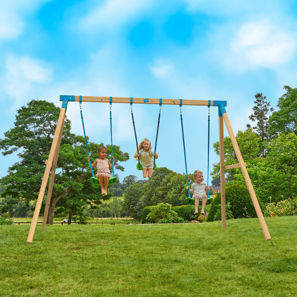 Three children swing on the TP Toys TP Kingswood Triple Squarewood Swing Set (FSC® certified) in a grassy outdoor area, surrounded by trees and blue sky.