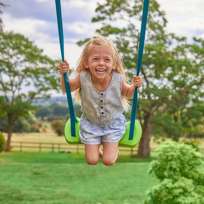 A young girl with blonde hair smiles while swinging outdoors on the TP Kingswood Triple Squarewood Swing Set by TP Toys, which features a green swing and sturdy wooden frame surrounded by grass and trees.