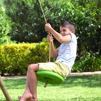 A young boy enjoys swinging barefoot on the TP Toys Sherwood Triple Wooden Swing Set (PEFC certified), smiling as he holds the rope and swings outdoors in the garden.