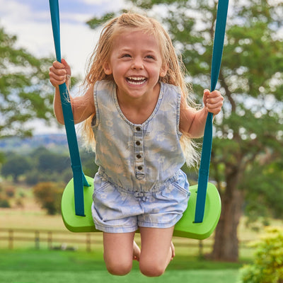 A young girl with long blonde hair smiles as she swings on the TP Sherwood Triple Wooden Swing Set by TP Toys, enjoying the outdoors amid trees and grass. PEFC certified.