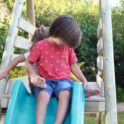 Two children sit together atop the TP Toys Knightswood Double Wooden Swing & Slide Set, with one child partially hidden behind the other, surrounded by greenery and the cheerful outdoor playset.